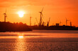 Industrial buildings along with wind turbines and dock cranes on a river harbour silhouetted against a fiery sky at sunset