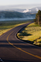 A Sunlit Road Winds Through Hayden Valley In Yellowstone National Park, Wyoming With Mist Rising In The Background.