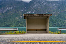 Gray Concrete Bus Stop On The Side Of The Road Behind Which There Is A Fjord With Blue Water And Behind The Fjord There Is A Beautiful View Of Big Mountains
