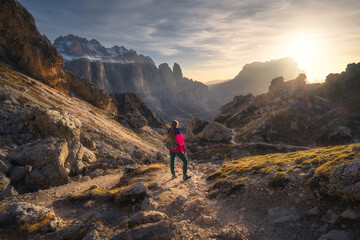 Wall Mural - Girl with backpack on the trail in mountains at sunset in autumn. Beautiful landscape with young woman, high rocks, path, stones, orange grass, sky with clouds in fall in Dolomites, Italy. Adventure