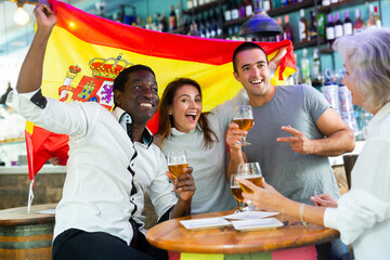 Wall Mural - Spainian football, soccer fans cheering goal their team with glasses of alcohol drink at pub, supporting favourite players, man showing victory sign. Concept of sport, human emotions