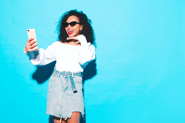 Wall Mural - Beautiful black woman with afro curls hairstyle. Smiling model dressed in white summer top and jeans clothes. Sexy carefree female posing near blue wall in studio. Tanned and cheerful. Takes selfie