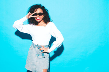 Wall Mural - Beautiful black woman with afro curls hairstyle. Smiling model dressed in white summer top and jeans clothes. Sexy carefree female posing near blue wall in studio. Tanned and cheerful. Isolated