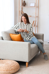 Canvas Print - Young woman with glass cup of coffee sitting on grey sofa at home