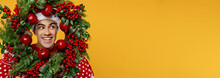 Close Up Merry Fun Young Man Wearing Red Knitted Sweater Santa Hat Posing Hold Look Aside Through Christmas Wreath Isolated On Plain Yellow Background Happy New Year 2023 Celebration Holiday Concept.