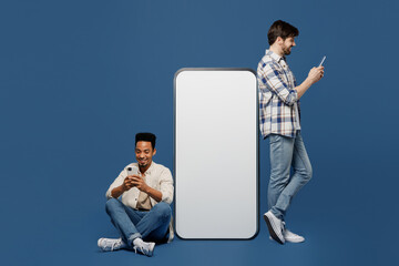 Wall Mural - Full body side view young two friend men wear casual shirts together sit near big huge blank screen mobile cell phone gadget with mockup use smartphone isolated plain dark royal navy blue background.