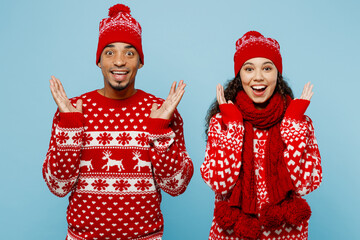 Merry young surprised shocked couple friends two man woman wear red Christmas sweater Santa hat posing spread hands isolated on plain pastel light blue background. Happy New Year 2023 holiday concept.