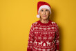 Happy confident senior woman wearing red santa hat stands and looking at the camera isolated on yellow. Mature lady promoting new year deal, holiday sale