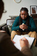 © AnnaStills - African American woman in depression sitting on sofa and talking to psychologist while he making notes in document