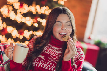 Photo of nice adorable sweet overjoyed girl with brunette hair wear red ornament jumper enjoy cookie hold tea cup in house indoors