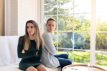 Dissapointed young couple turning their back on each other while sitting in the sofa in the living room with garden