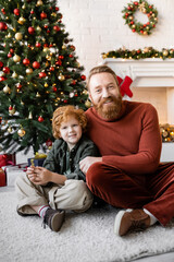 Wall Mural - cheerful father and child with red hair sitting on floor near Christmas tree and looking at camera