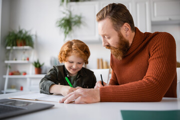 Wall Mural - smiling kid writing in notebook near bearded dad and laptop on blurred foreground