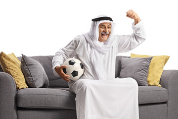 Wall Mural - Mature arab man in a robe holding a football and cheering seated on a sofa