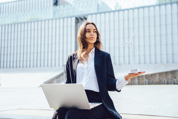 Wall Mural - Successful young woman with wind turbine model and laptop sitting outdoor with modern corporate building exterior.