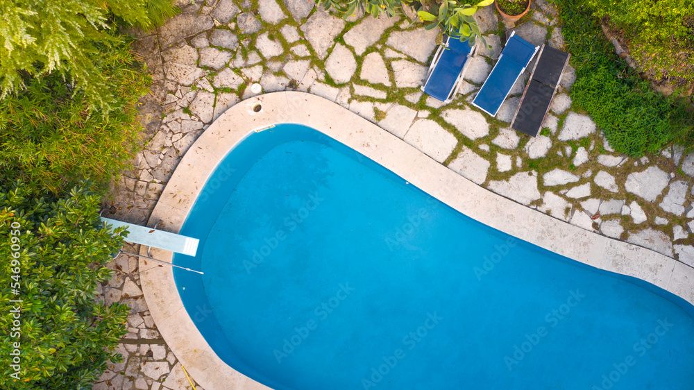 Aerial view of a circular swimming pool with diving board, belonging to a large villa. The water is transparent and blue. Around the water there is a stone floor and a garden.