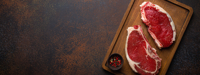 Wall Mural - Two raw uncooked meat beef rib eye marbled steaks on wooden cutting board with seasonings on dark rustic background ready to be grilled from above, preparing dinner with meat, space for text