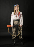 Fototapeta Sport - Full length portrait of a Ukrainian female soldier wearing a Ukrainian embroided shirt with a wreath and holding a rifle isolated on a black