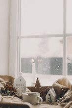 Warm Cup Of Tea, Candles, Lights, Little Christmas House And Tree, Wooden Star On Cozy Blanket On Windowsill. Winter Hygge. Atmospheric Christmas Background. Cozy Home On Snowy Day