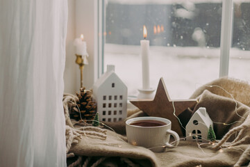 Wall Mural - Warm cup of tea, candles, lights, little christmas house and tree, wooden star on cozy blanket on windowsill. Winter hygge. Atmospheric Christmas background. Cozy home on snowy day