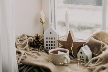 Wall Mural - Cozy home at snowy day. Warm cup of tea, candles, lights, little christmas houses and trees, wooden star on cozy blanket on windowsill. Winter hygge. Atmospheric scandinavian mood