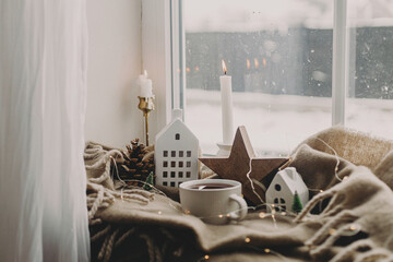 Wall Mural - Winter hygge. Warm cup of tea, candles, lights, little christmas houses and trees, wooden star on cozy blanket on windowsill. Cozy home. Atmospheric snowy day. Christmas background