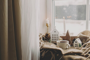 Wall Mural - Warm cup of tea, candles, lights, little christmas house and tree, wooden star on cozy blanket on windowsill. Winter hygge. Atmospheric Christmas background. Cozy home on snowy day