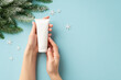 Winter skin care concept. First person top view photo of young woman's hands holding white tube without label snowflakes and pine branch in frost on isolated pastel blue background with empty space