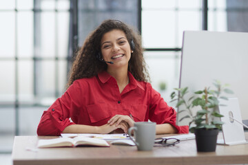 A young attractive woman in a red shirt sits at a table in a red jacket in headphones with a microphone.