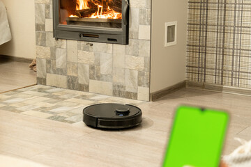 Smartphone screen with chromakey against cozy light room. Modern black robotic vacuum cleaner cleans tiled floor near fireplace in apartment closeup