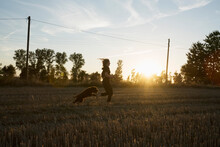 Carefree Girl And Dog Running In Sunny Rural Farm Field At Sunset

