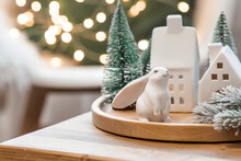 Beautiful Holiday Card. The Concept Of A Happy New Year And Christmas 2023. Christmas Trees, A White Rabbit And Scandinavian White Houses On A Wooden Table In A Cozy Living Room.