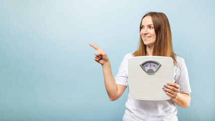 Woman with weighing scale pointing with finger to side on blue background