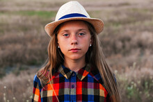 Defocus Girl Kid In Hat Looking At Camera. Portrait Of Sad Little Girl At The Day Time. Meadow Nature Background. Out Of Focus