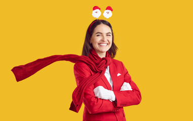 Beautiful brunette is smiling sweetly, dressed in a red business suit with Santa Claus figurines on her head and a red scarf. The model stands with her arms crossed on an isolated yellow background.