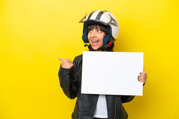 Wall Mural - Young latin woman with a motorcycle helmet isolated on yellow background holding an empty placard and pointing side