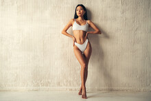 Full Length Of Asian Woman In White Bikini Posing Near Textured Wall