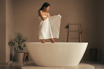 Rare view of attractive asian slim woman in towel staning in  white ceramic bathtub to refresh