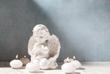 Little Statue Of White Angle And White Burning Candles Against Blue Textured Wall. Selective Focus.