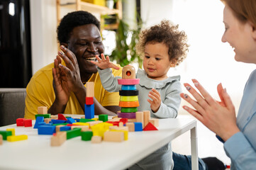 Wall Mural - Mother and father supporting their cute little daughter in playing with colorful didactic wooden toys at home