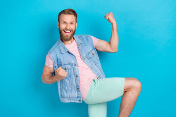 Poster - Photo of positive lucky young guy dressed denim vest smiling rising fists isolated blue color background