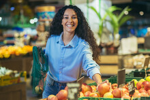 Portrait Of Happy Woman Shopper In Supermarket, Hispanic Woman Chooses Apples And Fruits Smiling And Looking At Camera, With Grocery Basket Chooses Goods.