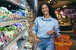 © Liubomir - Portrait of happy hispanic woman shopper in supermarket, woman with shopping basket smiling and looking at camera, near shelves with products in grocery department.