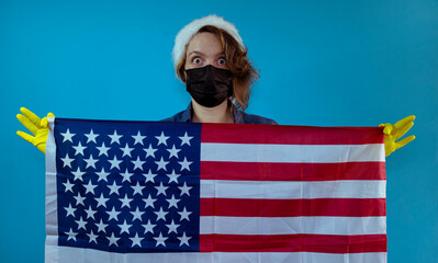 Wall Mural - An adult woman in a black mask and a New Year's hat holds a USA flag in her hands on an isolated blue background. American woman portrait. Flag day. Self isolation in the new year