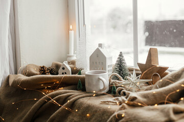 Wall Mural - Winter hygge, Christmas still life. Warm cup of tea, christmas decorations, lights, little house, star on cozy blanket on windowsill. Cozy home on snowy day. Atmospheric scandinavian mood