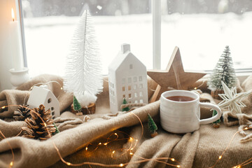 Wall Mural - Christmas still life, winter hygge home. Warm cup of tea, christmas decorations, lights, little house, star on cozy blanket on windowsill. Cozy home. Atmospheric scandinavian mood