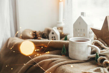 Wall Mural - Winter hygge, Christmas still life. Warm cup of tea, christmas decorations, lights, little house, star on cozy blanket on windowsill. Cozy home on snowy day. Atmospheric scandinavian mood