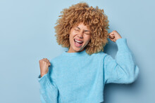 Excited Cheerful Woman With Curly Hair Clenches Fists Raises Arms Celebrates Success Exclaims From Joy In Casual Jumper Glad To Achieve Goal Isolated Over Blue Background. Victory Celebration