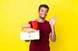 Young brazilian man holding pizzas and burgers isolated background saluting with hand with happy expression