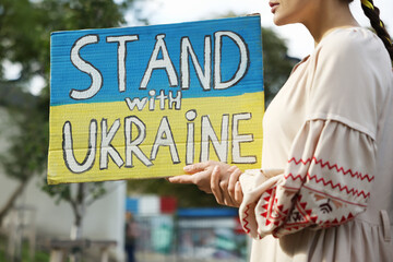 Wall Mural - Young woman holding poster in colors of national flag and words Stand with Ukraine outdoors, closeup